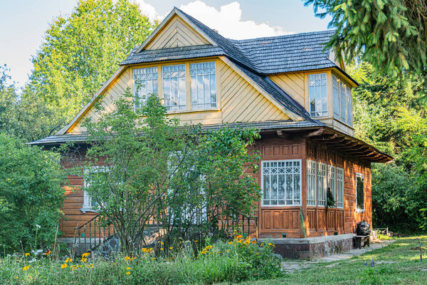 A two-story wooden house in a forested village, painted yellow and brown, with a small garden enclosed by a rustic fence No people are present, and the ample daylight suggests a tranquil setting