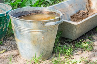 Eski bir Orta Avrupa köyünde dış mekan sahnesi. Çamurlu suyla dolu büyük metal kova, muhtemelen yakınlarda çamur içeren daha küçük yeşil plastik konteynırları karıştırmak için kullanılıyor.