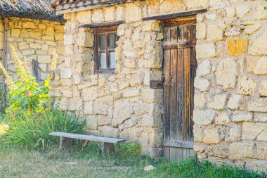 Orta Avrupa köyündeki eski taş kulübe düzensiz şekilli taşlar, ahşap kapı, çoklu cam pencere, yıpranmış kepenkler, boş bank, yamalı çimler, yumuşak ışık şafağı, doğal renkler.