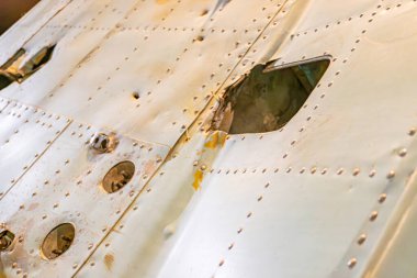 Korozyonu, görünür perçinleri, dairesel girintileri olan eski model bir uçak yapısının yakın çekimi ve üst merkezin metalik malzeme dokusunu açtığı açıkça görülüyor.