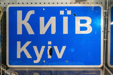 Kiril alfabesindeki hasar görmüş yol işareti, metal bir ızgara yapısına yaslanmış, kapalı alanda ya da açık havada kurşun delikleriyle kısmen gizlenmiş, hatta gizli fotoğraf tarzını bile ışıklandırmış.