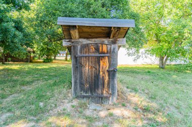 Orta Avrupa köyünün çimenli bölgesinde kırsal, yıpranmış bir tuvalet, bulutlu bir gün, seyrek bitki örtüsü, arka planda ağaçlar, hiç insan yok.
