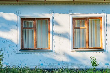 İki pencereli eski Orta Avrupa binası, biri kapalı, biri yarı kapalı balkon korkuluğu görünür açık renkli ön cephe, gündüz vakti aydınlatma işaretleri var.