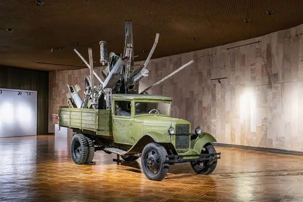 Eski, zeytin yeşili, jantlı ve haberleşme cihazlı bir kamyonet kapalı bir müze sergi alanında parlak, desenli bir zemine park edilmiştir.