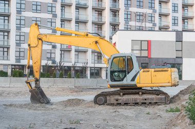 Sarı kazıcı bir inşaat sahnesine çapraz yerleştirilmiş, kovası arka planda yenilenmekte olan çok katlı gri bir binanın içine kazılmış, beyaz bir yapının bitişiğindeki Bulutlu Sek