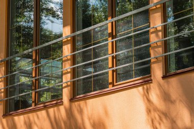 Modern bir binanın ön cephesinde portakallı çizgili bir duvardaki koyu çerçeveli pencerelerin dikey sırasına yakın çekim, gün ışığını ve bitki örtüsünü yansıtır.