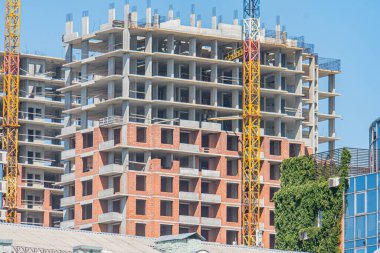 Sarı ve kırmızı vinçlerle zıt beton ve tuğla yapıların yer aldığı çok katlı bir inşaat alanı ön planda yeşil yaprakların bahar ya da yaz olduğunu gösteriyor.