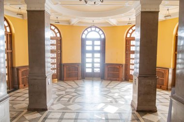 Eski bir Avrupa şehrinde sarı duvarlı büyük oda kemerli, beyaz perdeli desenli pencere, büyük ahşap masa, sıralı sandalyeler, desenli döşeme, çevre aydınlatması, karmaşık pencere camları