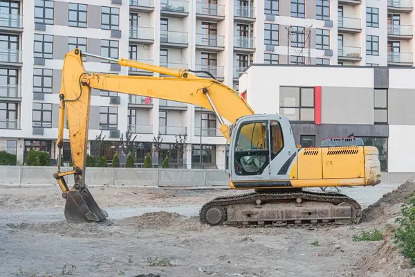 Sarı kazıcı bir inşaat sahnesine çapraz yerleştirilmiş, kovası arka planda yenilenmekte olan çok katlı gri bir binanın içine kazılmış, beyaz bir yapının bitişiğindeki Bulutlu Sek