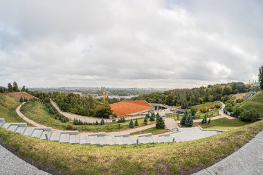 Dairesel amfi tiyatro, dolambaçlı yol, ağaçlar, bulutlu gökyüzü ve uzak binalarla dolu yemyeşil bir parkın panoramik manzarası