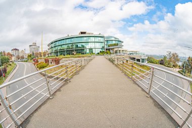 Gökyüzünü yansıtan kavisli modern cam bina, şehir arka planı, yaya yolu, durgun su, metal korkuluk, yeşillik, panoramik manzara