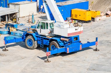 Büyük mavi vinç kamyonu, gri betona park etmiş, küçük su birikintileri var, kaportası aşınmış, kolu sarı kancaya takılmış, arka planda endüstriyel yapılar kamyonu, nakliye konteynırı,