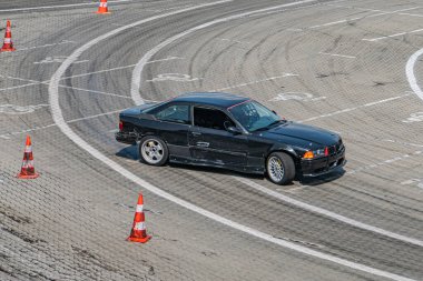 Gümüş jantlı siyah bir spor arabanın dinamik görüntüsü, turuncu kozalaklı gri bir yarış pistine bakıyor. Araba, son kullanılan doğal ışıklandırma kalıplarından aerodinamik değişiklikler ve lastik izleri gösteriyor.