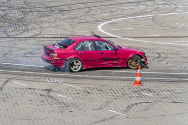 Canlı kırmızı bir yarış arabası, solda turuncu bir koni bulunan havadan bakıldığında gri bir pistte ilerliyor. Görüntü, yumuşak gölgeler ve doğal ışıklandırmayı betimliyor.