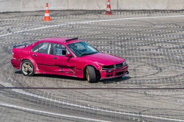 Kırmızı, ağır modifiye edilmiş bir sedan, beyaz yarış çizgileri olan bir yarış pistinin ortasında yakalanır. Araba, geniş çaplı motor sporlarından kalma aşınma ve yırtık izleri gösterir.