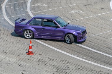 Yarış pistindeki dinamik bir sahnede mor bir BMW E36, plaka B36, keskin tekerlek açısı ve vücut eğimine sahip dar bir sola doğru eğimli turuncu bir trafik konisi rayların kenarını işaretliyor.