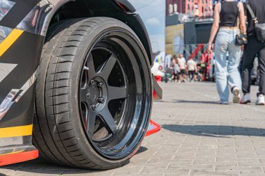 Siyah ve kırmızı renkli lastik, parlak tekerlekli, birden fazla desenli, yandan okunamayan metin yan lastikte, dekore edilmiş araba kaportasına karşı kısmen görülebilir, muhtemelen markalaşma ya da sponsorluk...