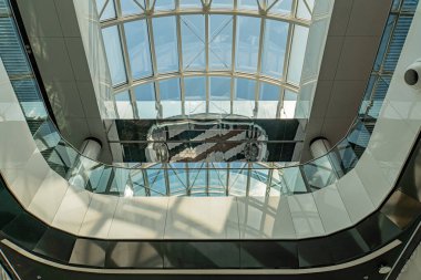 Yüksek tavanlı modern atriyum, büyük çatı pencereleri, yansıtıcı duvar panelleri, kıvrımlı cam merdiven, sanat eseri, minimalist tasarım