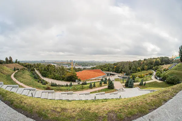 Dairesel amfi tiyatro, dolambaçlı yol, ağaçlar, bulutlu gökyüzü ve uzak binalarla dolu yemyeşil bir parkın panoramik manzarası