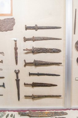 Bir müzede sergilenen antika Roma kılıçlarının koleksiyonu, bej bir duvardaki kancalara asılmış, pas ve aşınmayla düzenlenmiş, farklı dönemleri gösteren çeşitli tasarımlar.