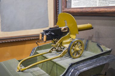 Müze ortamında eski model ateşli silah, koyu ahşap döşeme, mat metal parçalar, sarı dekoratif panel, çerçeveli belgeler, hatta ışıklandırma.