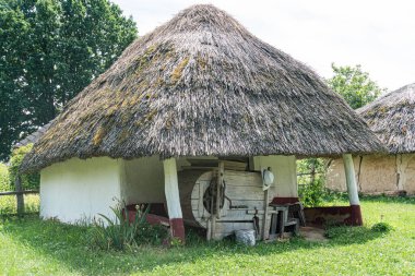 Eski Orta Avrupa 'da sazdan çatılı bir kulübede, muhtemelen tarım için kullanılıyor, kırsal bir alanda tarım makineleri, enkaz parçaları, ve çevredeki dünyevi renk paleti, görülebilir insan yok, açı!