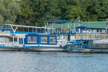 Huzurlu bir rıhtımda demirlemiş iki konut teknesi var. Birinin güvertesi mavi, diğerinin ise kapalı bir kabini var. Rıhtım yemyeşil sulara kadar uzanıyor.