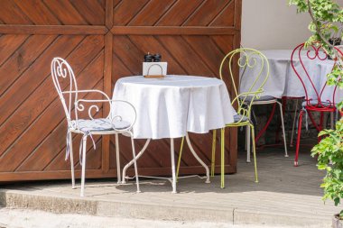 Modern bir şehir terası sahnesinin yakın çekimi Demir masa, beyaz bez, yeşil sarı sandalyeler, gün ışığı, yumuşak gölgeler, ahşap yapıya yakın gerçekçi yemek düzeni.