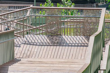 Ahşap köprü, yumuşak gölgeler ve berrak gökyüzü ile karakterize modern bir şehir sahnesinde, sakin bir su kütlesinin üzerinde zarif bir şekilde kavisli bir şekilde uzanır.