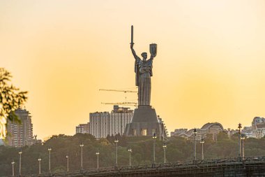 Heybetli heykel uzun figür, yükseltilmiş kol, Kyivs şehrinin siluetinde potansiyel bir nesne, muhtemelen günbatımında günbatımında su kütlesi renkleri, ağaçlar, kentsel binalar, yüksek binalar, inşaat işaretleri yansıtıyor.,