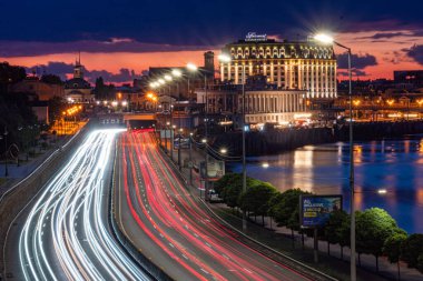 Ukrayna 'nın Ukrayna otoyolunun beyaz ve kırmızı ışıktan oluşan dinamik şablonlar oluşturduğu Nightscape of Kyiv geniş, aydınlık bina bir odak noktası olarak hizmet veriyor Su B. Kyiv, Ukrayna 06-29-2025