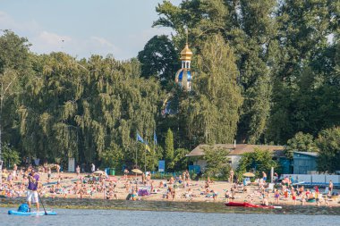 Kyivler 'in sakin sahili üzerinde açık gökyüzü, uzanan insanlarla dolu, sörf yapan veya kano oynayan yemyeşil ağaçlar, belirgin mavi kubbeli bui Kyiv, Ukrayna 07-12-2025