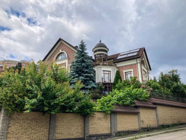 İki katlı kırmızı ve beyaz konut binası bacalı, açık merdivenli, girişi kapalı, balkon, yemyeşil bahçe, yeşil ağaçlar, ahşap çit, bulutlu gökyüzü, doğal gün ışığı.