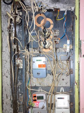 Çeşitli bileşenler, kablolar, devre kesiciler, anahtarlar ve metre içeren sarı duvarlı bir elektrik panelinin yakın görüntüsü. Merkezi metre kutusu metal elementlerle çelişiyor.