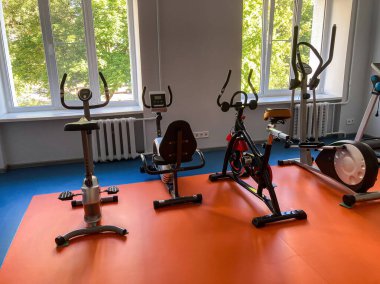 Üç egzersiz makinesinin, iki koşu bandının ve bir eliptik eğitmenin olduğu iki pencerenin doğal ışığına karşı iyi ışıklandırılmış bir spor salonu.