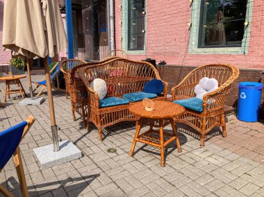 Açık kafe seti, hasır mobilyalar, bank, mavi yastıklar, cam masa, asfaltlı fayanslar, yeşil ipuçları, şemsiye serin atmosfer, parlak, hatta aydınlatma, muhtemelen sabah erken ya da geç