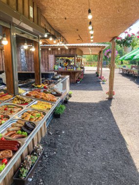 Hareketli, hareketli bir yiyecek standı, kıvrımlı metal bir çatı altında, çeşitli yiyecekler ve belirgin bir ızgara sergiliyor. Çevrede saksı bitkileri ve arka plandaki diğer yapılar var. Doğal da