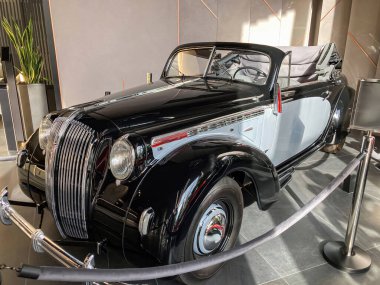 1930 'lardan kalma parlak, koyu renkli eski bir araba sergi salonunda sergilenir. Arka planda ince ışıklandırmalı ve benzer bir araba daha görünür. Arabanın krom kaplaması var.