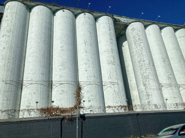 Açık gökyüzünün altındaki açık sanayi bölgesinde büyük, beton ve beyaz silolardan oluşan bir grup silo yüzeyleri ve bitki örtülerini tabanlarındaki alçak bir bariyerde büyüttüler.