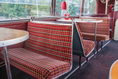 Klasik Londra çift katlı otobüs içi, ılık renk şeması kırmızı, turuncu, sarı, retro desenli koltuklar, dairesel metal aydınlatma armatürü, yüksek perspektif, doğrusal yol koridoru