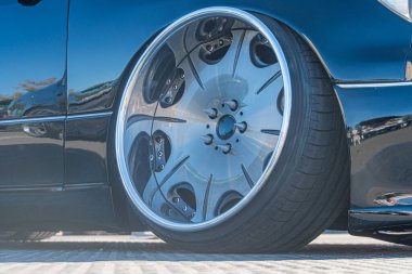 Arka planda gümüş ya da krom kaplama, siyah lastik ve koyu renk araba kaportası olan, dörtte üç açılı bir araba tekerleğinin yakın görüntüsü muhtemelen asfalt ya da beton zemindir.