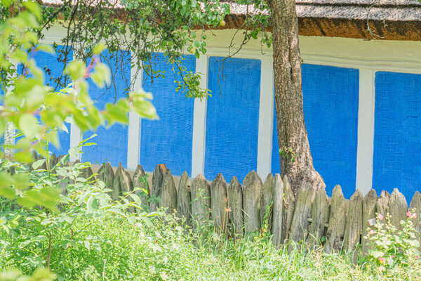 Частично видимый синий дом с белой отделкой на заднем плане, обрамленный пышной зеленью Старый деревянный забор, ветхий, но деревенский, на переднем плане E