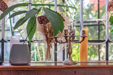 Pencere pervazında süs eşyası koleksiyonu, saksı bitkisi, metal tutacaklarda mumlar, soyut seramik heykel yumuşak gölgeler, dengeli kompozisyon, doğal ışık