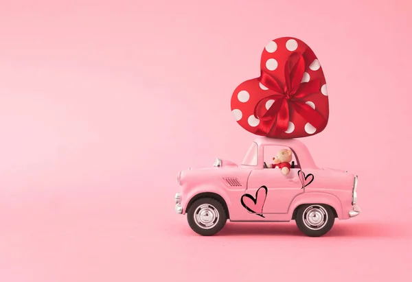 Love composition made of Pink toy car delivering gift box in the shape of a heart with ribbon and bow on pink background. Minimal concept of Valentine's Day or love. Creative art, minimal aesthetics.