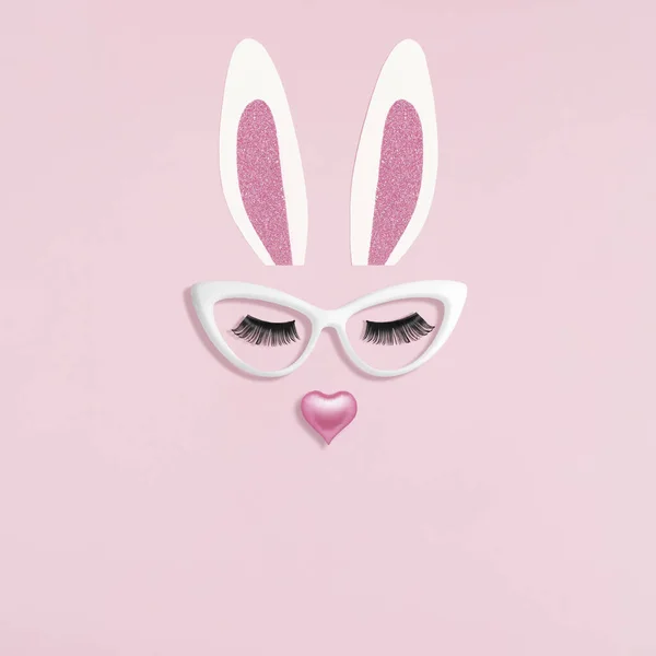 Bunny rabbit face made of cardboard ears, artificial eyelashes with glasses and pink heart on pastel pink background. Flat lay. Happy Easter minimal concept