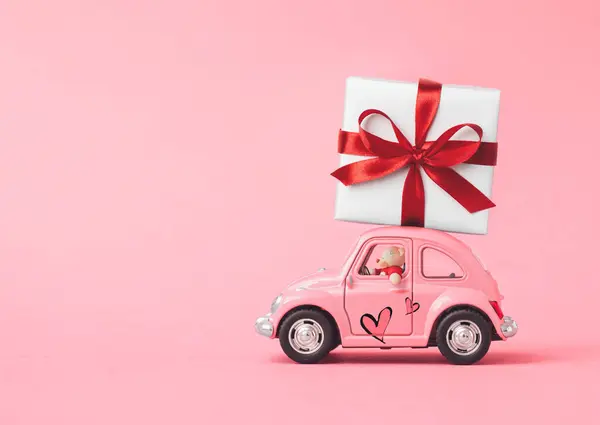 Love composition made of Pink toy car delivering gift box with ribbon and bow on pink background. Minimal concept of Valentine\'s Day or love. Creative art, minimal aesthetics.