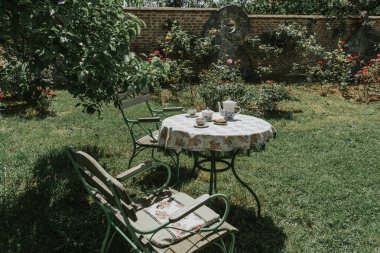 Çiçek açan güller ve yeşillikler arasında çay masası olan büyüleyici bir bahçe görüntüsü sükunet ve rahatlığı çağrıştırıyor..
