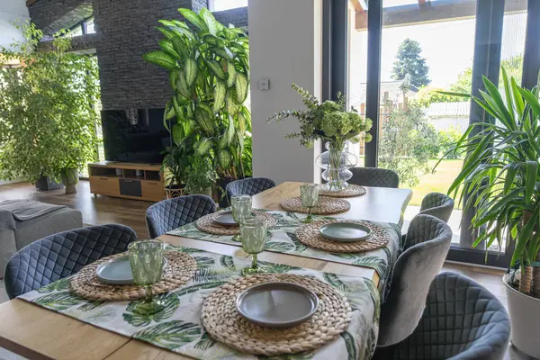 Geniş pencerelerden güneş ışığıyla aydınlatılan, yeşil bitkiler ve ekolojik esinlenilmiş iç tasarım ve doğal elementleri vurgulayan zevkli sofra eşyalarıyla süslenmiş şık bir iç mekan. .