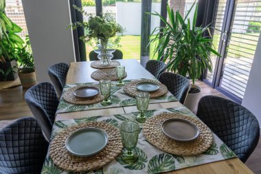 Geniş pencerelerden güneş ışığıyla aydınlatılan, yeşil bitkiler ve ekolojik esinlenilmiş iç tasarım ve doğal elementleri vurgulayan zevkli sofra eşyalarıyla süslenmiş şık bir iç mekan. .