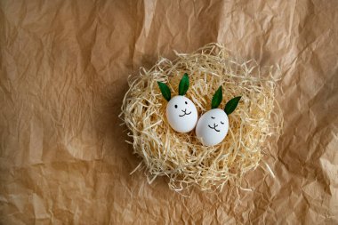 Fancy white Easter eggs with funny green bunny ears in straw nest on brown paper background. Flat lay, top view. Traditional spring concept.